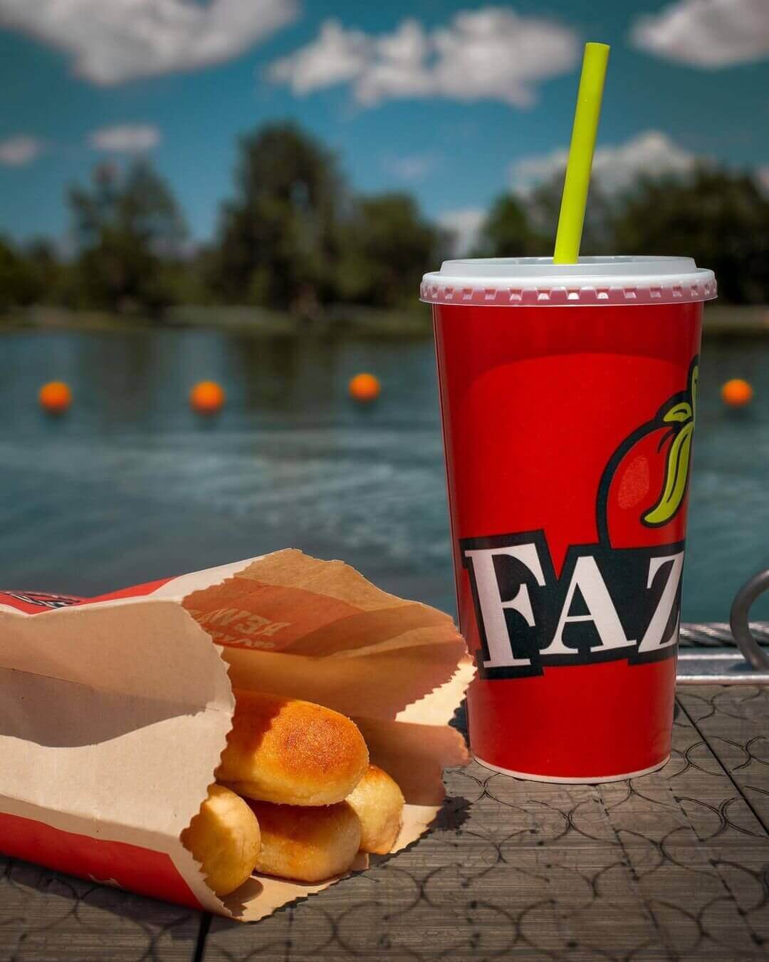 Bag of breadsticks and red to-go cup filled with soda sitting on a dock in front of a lake 