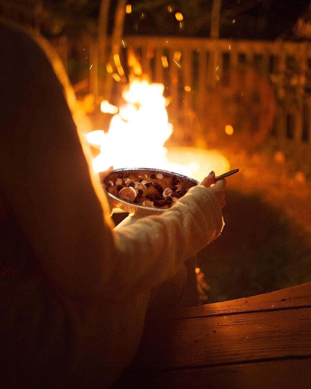 Woman enjoying s'mores breadstick bites out of aluminum container in front of flickering campfire at night 
