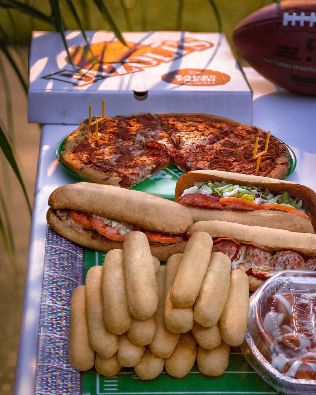 Tailgate table topped with a pizza, two sub sandwiches, breadsticks and a football