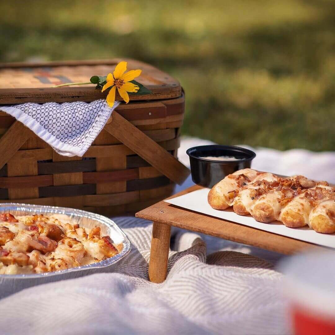 Picnic basket with a yellow flower sits behind two small trays of Italian food on picnic blanket 