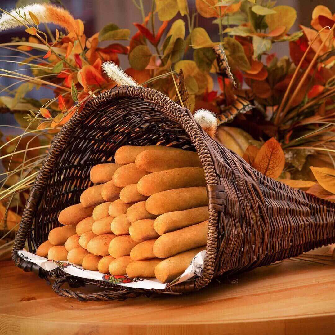 A large wicker cornucopia containing dozens of freshly baked breadsticks sits on a table in front of a bouquet of orange and yellow autum flowers and leaves  