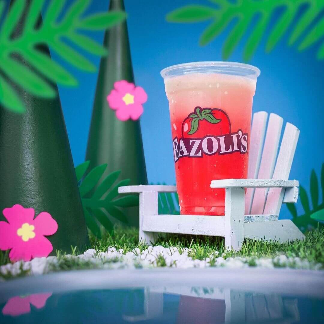 Diorama-style photo of a red slushie in a clear plastic cup on a tiny white beach chair in a stylized miniature beach scene 