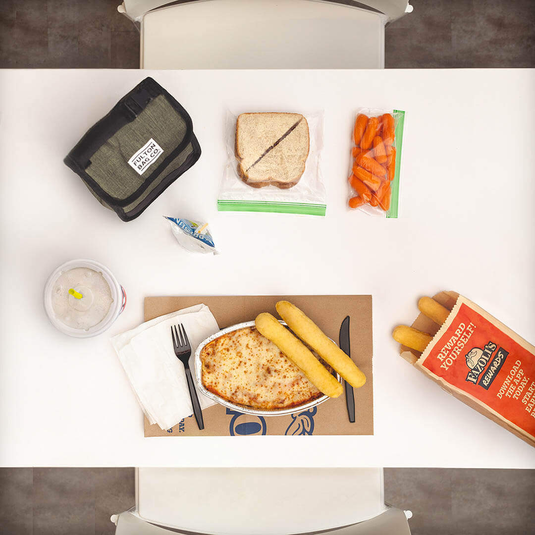 Overhead photo of a lunch table with a nicely arranged carryout dinner across from a traditional lunch with items like a PB&J  