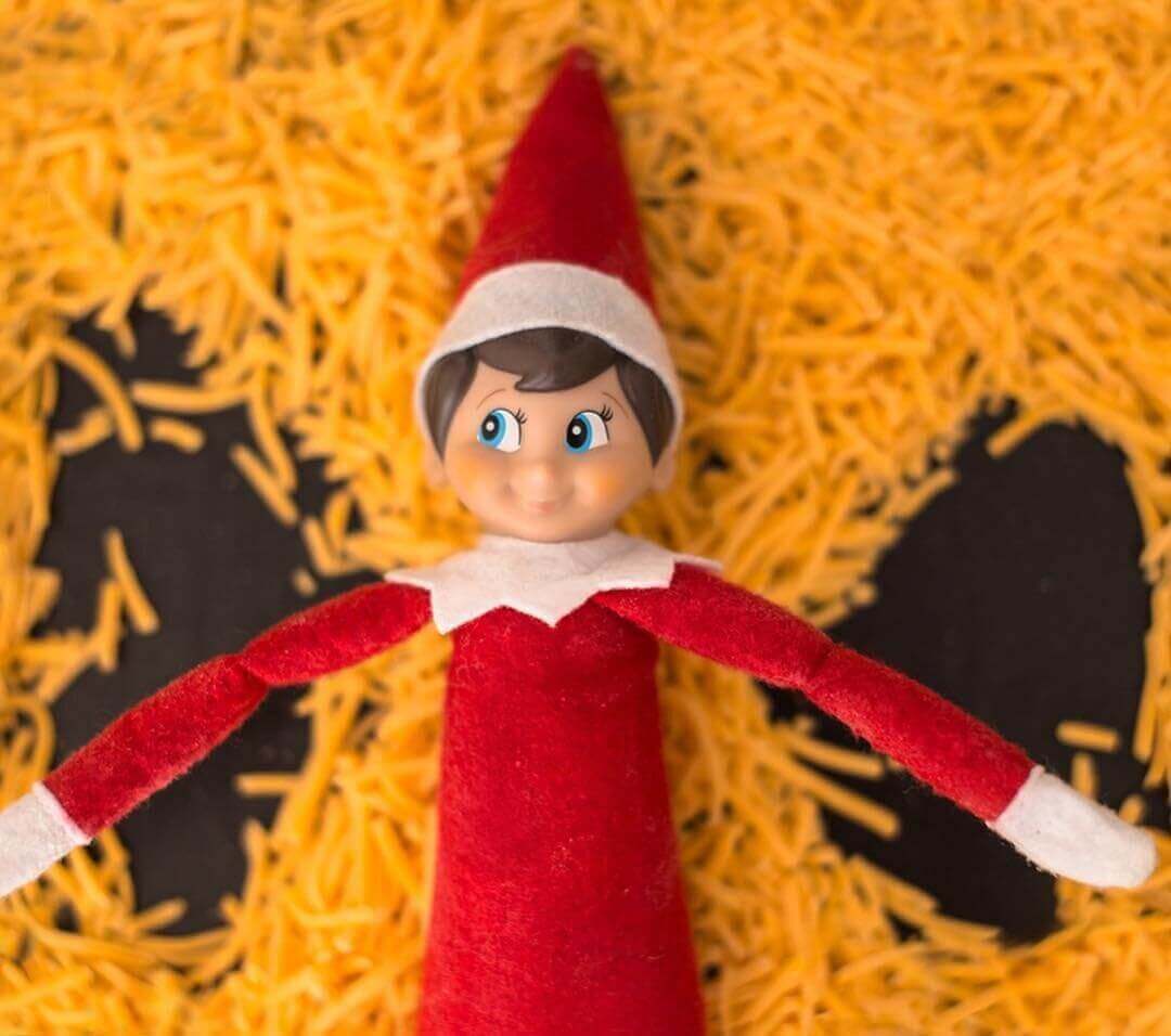 Overhead close-up photo of Elf-on-the-Shelf making snow angel in a pile of cheddar cheese 