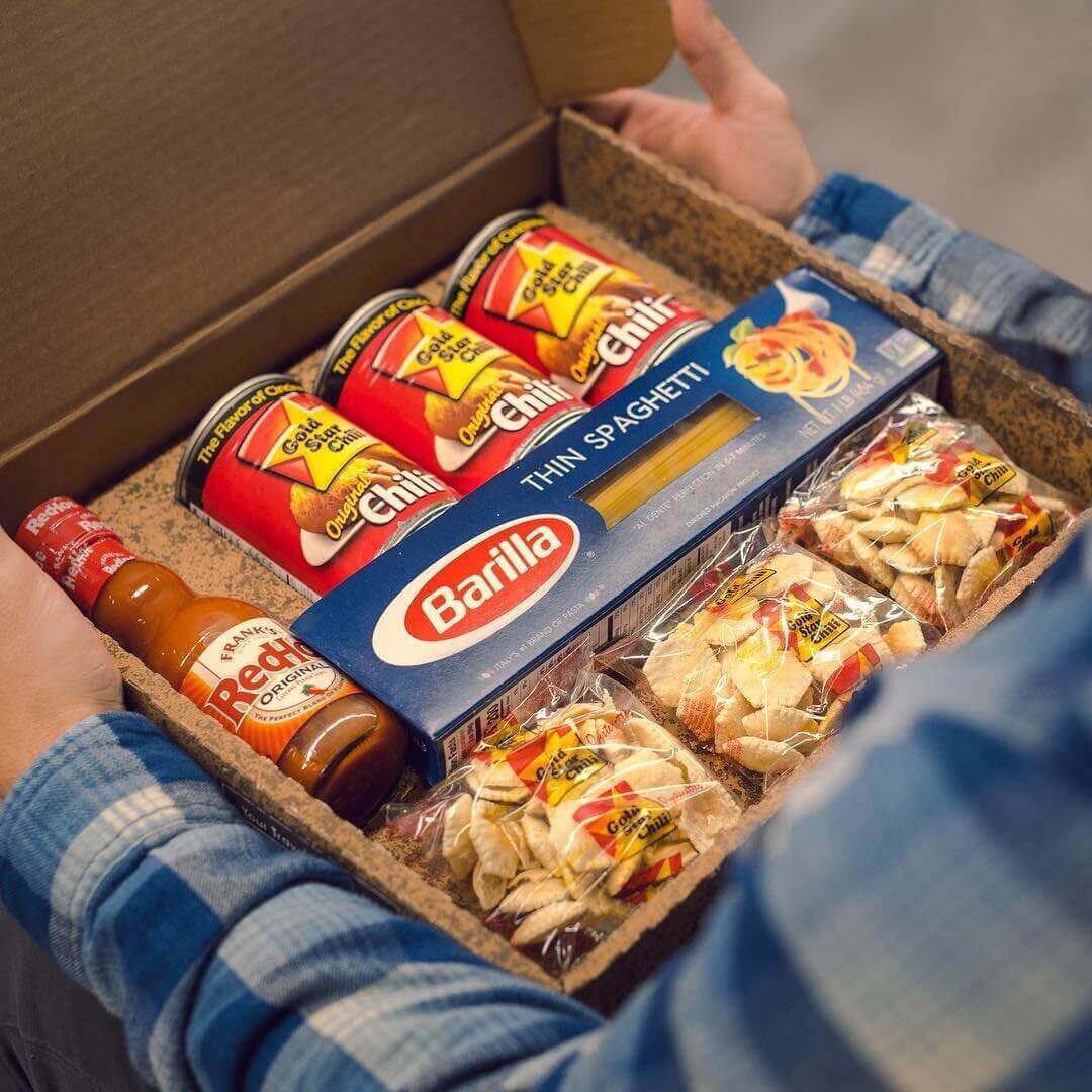 Over-the-shoulder photo of an open care package filled with cans of chili, boxed spaghetti, oyster crackers and a bottle of hot sauce 