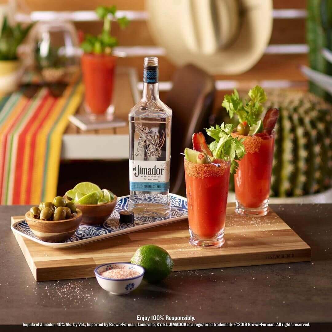Two bloody maria cocktails sitting on a cutting board next to a bottle of tequila and sliced limes 